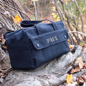May include: A black canvas tool bag with a top handle and a front flap secured by two black snaps. The name "PAUL" is printed on the front flap. The bag is sitting on a tree trunk with fall leaves.