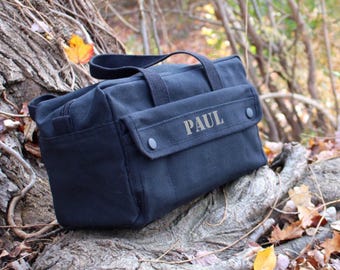 May include: A black canvas tool bag with a top handle and a front flap secured by two black snaps. The name "PAUL" is printed on the front flap. The bag is sitting on a tree trunk with fall leaves.