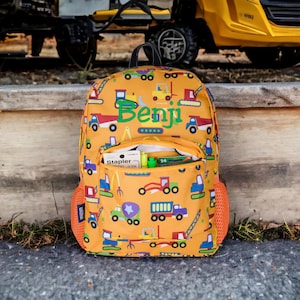 May include: A yellow construction vehicle backpack with a front pocket and a mesh side pocket. The backpack has a personalized name, "Benji", embroidered on the front.