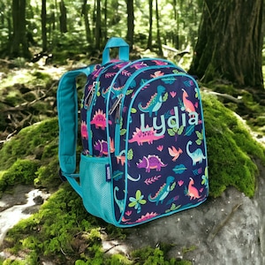 May include: A blue backpack with a dinosaur print and the name "Lydia" embroidered on the front. The backpack has a turquoise zipper and a mesh pocket on the side.