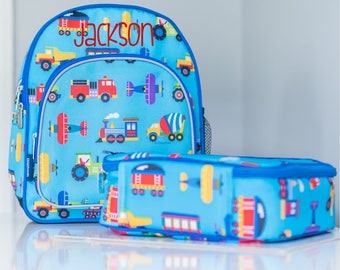 May include: A blue and red backpack with a lunch bag. The backpack has a blue background with a pattern of airplanes, trucks, and trains. The lunch bag has the same pattern. The backpack has the name "Jackson" embroidered on the front.