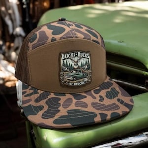 May include: A camouflage trucker hat with a brown front panel and a mesh back. The hat features a patch with the text "DUCKS-BUCKS N' TRUCKS" and an illustration of a deer, trees, and a truck. The brim has a camouflage pattern.