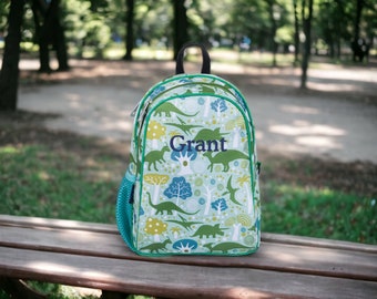 May include: A green and blue backpack with a dinosaur pattern. The backpack has a black zipper and a green trim. The name "Grant" is embroidered on the front of the backpack.