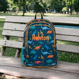 May include: A blue backpack with a dinosaur print and yellow accents. The backpack has a zippered pocket on the side and the name "Ashton" embroidered on the front.
