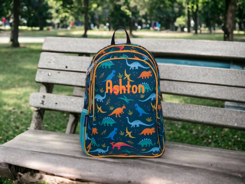 May include: A blue backpack with a dinosaur print and yellow accents. The backpack has a zippered pocket on the side and the name "Ashton" embroidered on the front.