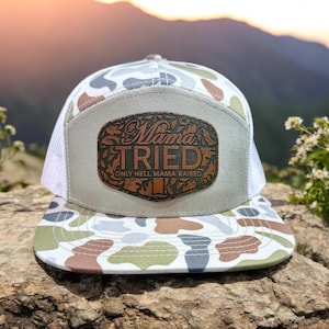 May include: A trucker hat with a camouflage pattern on the front and a white mesh back. The hat features a brown leather patch with the words "Mama Tried" and "Only Hell Mama Raised" in a decorative font.