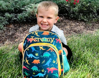 May include: A young boy smiles while holding a blue and green dinosaur backpack with the name "Jeremy" embroidered in yellow on the front.