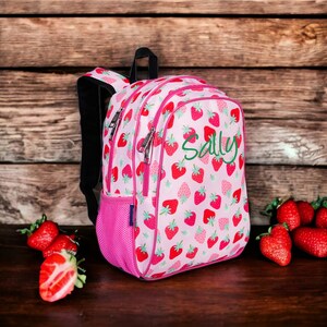 May include: Pink backpack with a strawberry pattern and the name "Sally" embroidered on the front. The backpack is sitting on a wooden surface with strawberries scattered around it.