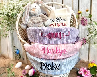 May include: A white wicker basket filled with Easter gifts. The basket is lined with three fabric liners, each with a different color and a name embroidered on it: Emma, Harlyn, and Blake. The basket also contains a stuffed bunny, a white towel, and a small bag with the text "He Miss Sorry I Spit Up..."