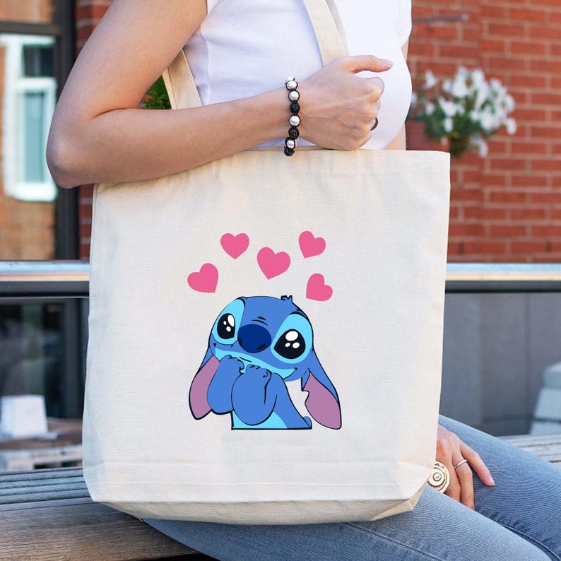 May include: Cream-colored tote bag featuring a blue cartoon character with large eyes and pink ears, surrounded by pink hearts. The bag has a simple design with a rectangular shape and long straps.