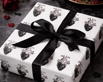 May include: A wrapped gift box with a black satin ribbon and bow. The wrapping paper is white with a repeating pattern of anatomical hearts in black. The scene includes red roses, candles, and heart-shaped confetti, creating a gothic aesthetic.