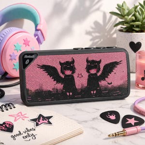 May include: A black Bluetooth speaker with a pink mesh front featuring a black graphic of winged figures. The speaker is surrounded by a pink and purple headset, a succulent plant, a candle, and stickers. The notebook reads "good vibes only."