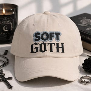 May include: A beige baseball cap with the words "SOFT GOTH" embroidered in black with a light blue outline. The cap has a curved brim and a button on top. The image also includes a black candle, a book, and gothic jewelry.