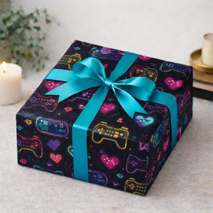 May include: A wrapped gift box featuring a black background with a pattern of colorful video game controllers and hearts. The gift is tied with a bright teal ribbon and bow, creating a festive presentation.