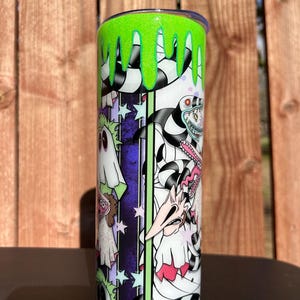 Könnte beinhalten: Ein zylindrischer Becher mit einem grünen Glitzerdeckel und einem tropfenden Schleimdesign. Der Becher hat ein Cartoon-Design mit schwarz-weißen Streifen, lila Akzenten und verschiedenen Charakteren.