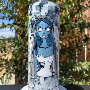 May include: A tall, cylindrical tumbler with a swirling white and gray pattern. The tumbler features a cartoon illustration of a blue-skinned woman in a white wedding dress and veil. The design is reminiscent of the character Emily from the movie "Corpse Bride".