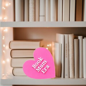 May include: A pink heart-shaped sign with the text "Book Mum Era" in white letters. The sign is sitting on a white bookshelf with books behind it.