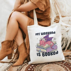 May include: A white tote bag featuring the text "MY WEEKEND IS BOOKED" and a cartoon dragon sleeping on books. The dragon is pink with purple wings. The bag is held by a person wearing brown boots.