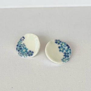 May include: A pair of white ceramic earrings with a blue floral design. The earrings are oval shaped and have a white background with blue flowers.