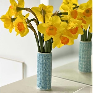 Könnte beinhalten: Ein Strauß leuchtend gelber Narzissen mit orangefarbenen Zentren in einer hellblauen, gemusterten Vase. Die Vase ist zylindrisch und hat ein strukturiertes Design. Die Blumen sind frisch und lebendig und bilden eine fröhliche Darstellung.