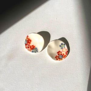 May include: A pair of white ceramic earrings with a floral design. The flowers are red, blue, and orange.