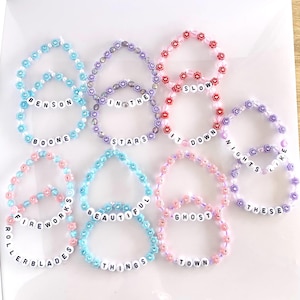 May include: A collection of 10 colorful beaded bracelets with different phrases on them, including "Benson", "Boone", "In the Stars", "It Down", "Nights Like These", "Fireworks", "Beautiful Things", "Ghost Town", "Rollerblades", and "Slow".
