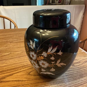 May include: A black ceramic ginger jar with a matching lid, featuring a hand-painted floral design in white, light blue, and gold metallic accents. The jar has a smooth, glossy finish and a rounded, bulbous body shape with a tapered base.