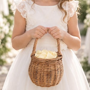 May include: A round, woven brown wicker basket with a sturdy handle, filled with soft cream-coloured flower petals. The basket features a classic, textured weave pattern and is held by a person wearing a white lace-trimmed dress with flutter sleeves.