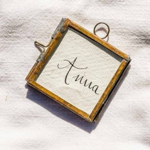 May include: A small, square, gold-toned frame with a glass front. The frame is hanging from a metal ring and has a clip on the side. The word "Anna" is written in black ink on a white background inside the frame.