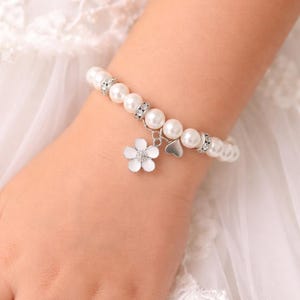 May include: A white pearl bracelet with silver detailing and charms. The bracelet has round pearls, silver spacers, and two charms: a white flower and a small heart. The bracelet is on a wrist, with a white lace background.