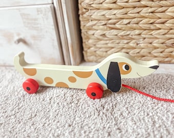 May include: A wooden pull-along toy dog with a cream body, brown spots, and red wheels. The dog has a black ear, a blue collar, and a red pull string. A classic toy for toddlers.
