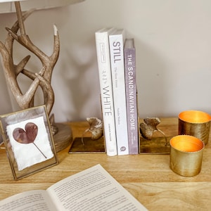 May include: A decorative display with books, leaf-shaped bookends, and candles. The books are titled "WHITE", "STILL The Slow Home", and "THE SCANDINAVIAN HOME". Two gold-coloured candles are also present. The bookends have a leaf design.