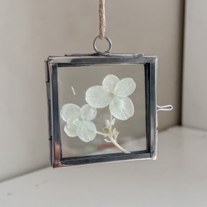 May include: A silver metal picture frame with a white pressed flower inside. The frame has a small loop at the top for hanging.