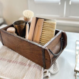 May include: A rectangular wooden box with dark metal handles and reinforced edges containing several cleaning tools. Inside are two wooden-handled brushes with light-coloured bristles, a round wooden scrubbing brush, and a dark metallic cup.