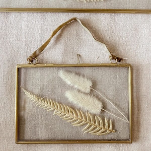May include: A gold-toned picture frame with a hanging ribbon. The frame holds dried white pampas grass and a dried fern.