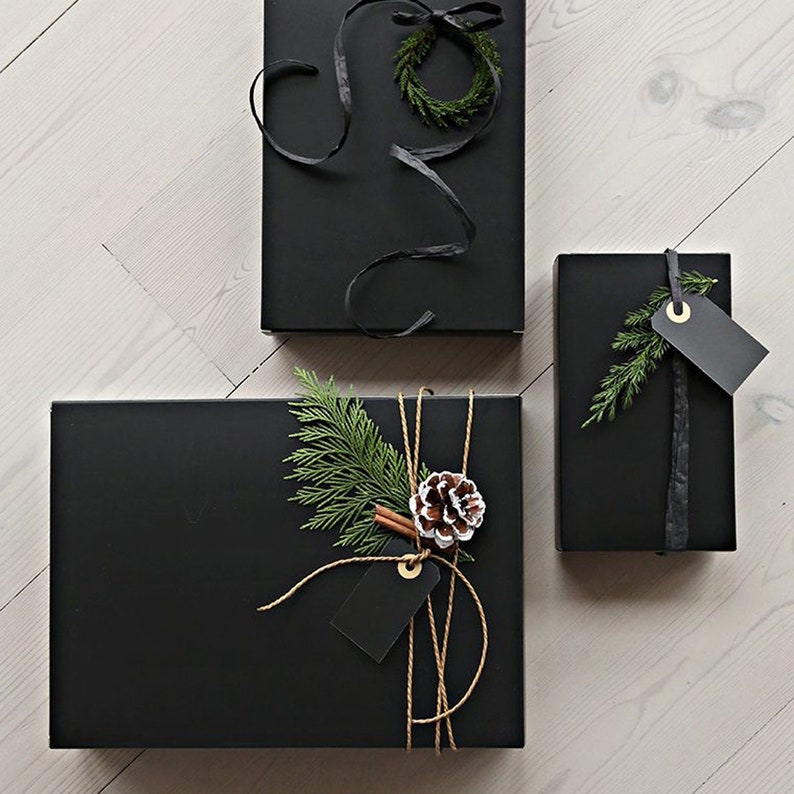 May include: Three black gift boxes wrapped with different decorations. One box has a small green wreath, another has a sprig of greenery and a pine cone, and the third has a sprig of greenery and a black tag.