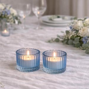 May include: Two lit blue glass votive candle holders with a ribbed design. The candles are on a white tablecloth, with flowers and glassware in the background. The votives have a soft, warm glow.