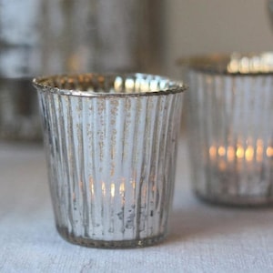 May include: Silver votive candle holders with a ribbed design. The holders are made of glass and have a clear, reflective finish.