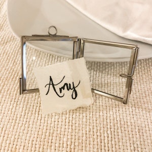 May include: A silver metal picture frame with a glass front and a hinged back. The frame is open and a small white card with the name "Amy" written in black ink is inside.