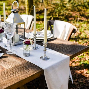 May include: A white linen table runner with silver candle holders and white candles on a rustic wooden table. The table is set with a glass of water and a small vase of flowers.