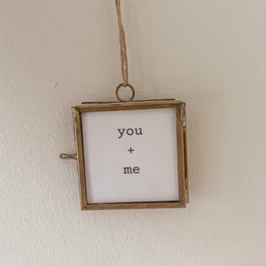 May include: A small, square, brass-toned picture frame with the words "you + me" printed on a white background. The frame is suspended by a thin, natural-colored cord. The frame has a clasp on the side.