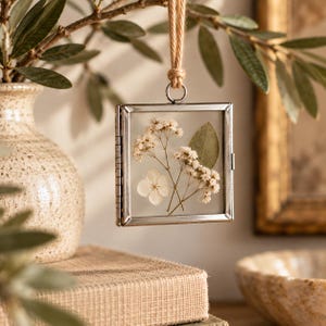 May include: A square, silver-toned metal floating frame hangs from a jute twine loop. Inside the two glass panes, there are pressed white baby's breath flowers, a single white hydrangea petal, and a small green leaf arranged centrally.