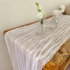 May include: White gauze table runner with a delicate, crinkled texture. Two clear glass vases with white flowers are placed on the runner.