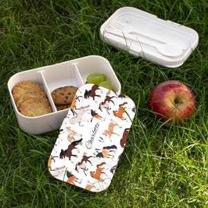 Op de afbeelding: Witte lunchbox met een deksel met een paardenpatroon en de naam "Charlotte". De doos is open en onthult koekjes, druiven en een koekje. Een rode appel en een apart compartiment met een vork en lepel zijn ook zichtbaar.