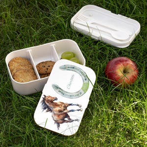 Op de afbeelding: Witte lunchbox met een deksel met een aquarel paard en hoefijzer ontwerp. De doos is open en onthult koekjes, druiven en een compartiment. Een rode appel en een apart deksel met bestek zijn ook zichtbaar.