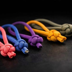 May include: A close-up shot of five brightly coloured paracord zipper pulls. The pulls are in shades of red, blue, purple, yellow, and olive green, each with a knotted design and a loop. Ideal for bags or clothing.