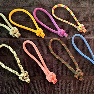 May include: Ten colorful paracord keychains with different knot designs. The keychains are made of various colors, including yellow, purple, pink, blue, and black.