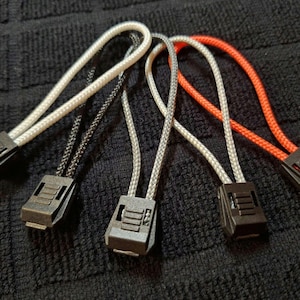 May include: Five zipper pulls in different colours, including black, white, grey, and orange. The zipper pulls are made of cord and have a plastic slider.