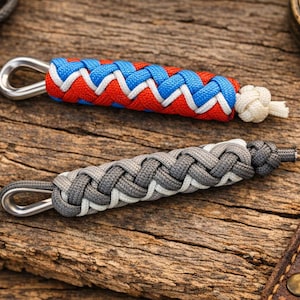 May include: Two paracord keychains with metal loops rest on a wooden surface. One is red, white, and blue, the other is gray and white. A compass and leather item are in the background.
