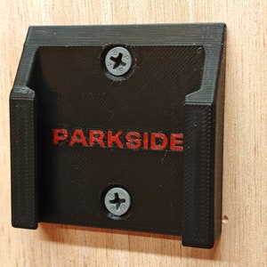 Peut inclure: Support mural en plastique noir avec le texte rouge "PARKSIDE". Le support est fixé à une surface en bois avec deux vis. Le support a une forme rectangulaire avec deux supports latéraux.